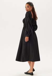 Zwarte jurk met lange pofmouwen, opgehaalde taille en vloeiende rok. Gladde textuur met een middellange uitvoering. Gecombineerd met eenvoudige zwarte ballerina's.