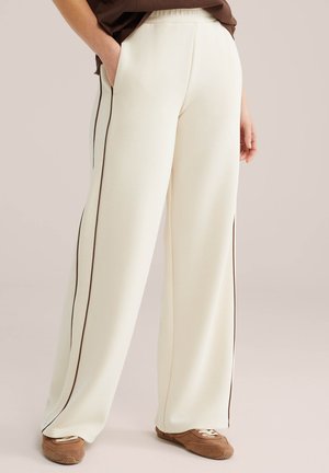 Personne portant un pantalon ample crème avec des bandes latérales marron et des baskets marron, debout avec une main dans la poche.