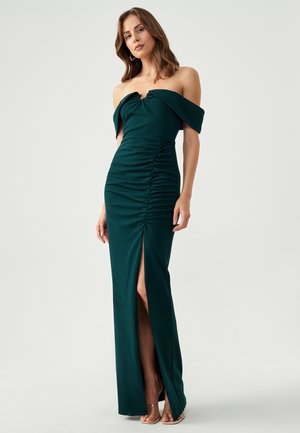 Green off-the-shoulder gown made of stretchy fabric. Features ruched detailing and a thigh-high slit on one side. Smooth texture.