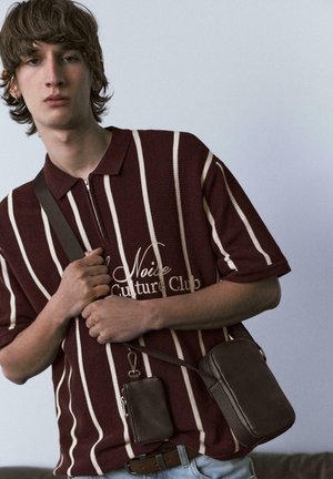 Młoda osoba ubrana w polo w bordowo-białe paski z napisem, trzymająca brązową torbę na ramię i małą saszetkę na jednolitym tle.