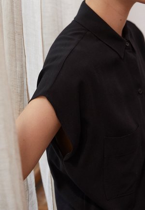 Zwarte kortek longsleeve shirt met een kraag, voorzien van een voorzak en een ontspannen pasvorm. Het materiaal lijkt glad met een matte textuur.