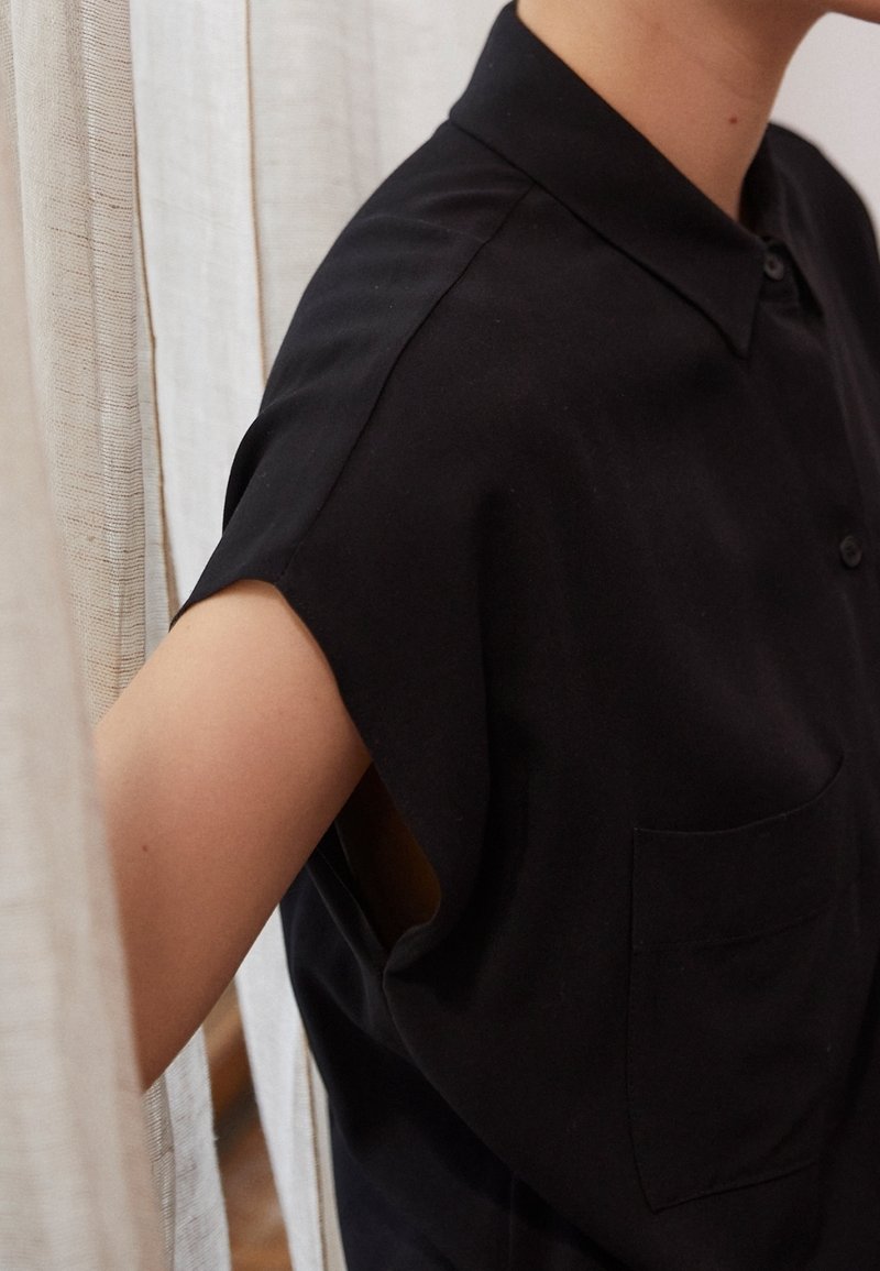 Camicetta nera a maniche corte con colletto, dotata di taschino frontale e vestibilità comoda. Il materiale sembra liscio con una texture opaca.