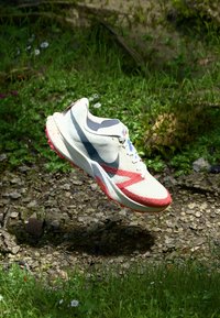 Běžecká obuv Nike, která má bílou síťovinovou svrchní část, modré a červené akcenty, texturovanou červenou podešev a výrazný design pro výkon.