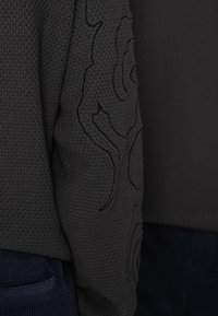 Dzianinowy ciemnoszary sweter o fakturowanej powierzchni z haftowanym wzorem na rękawie. Zaokrąglony dół i prążkowane mankiety.