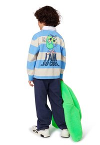 Ριγέ μακρυμάνικο πόλο μπλουζάκι σε μπλε και μπεζ αποχρώσεις, με μια καρτούν φιγούρα που φορά γυαλιά ηλίου στην πλάτη. Σκοτεινά παντελόνια και πράσινη φλις.