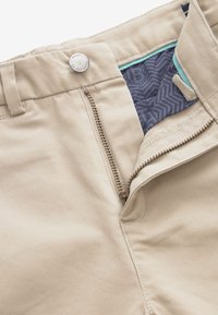 Beige broek met ritssluiting, metalen knoopsluiting en een gestreepte blauwe binnenvoering met een vissengraatpatroon. Glad stofoppervlak.