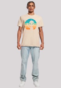 Beige cotton t-shirt featuring a distressed alien graphic, with blue, green, and orange horizontal stripes inside a circular design.