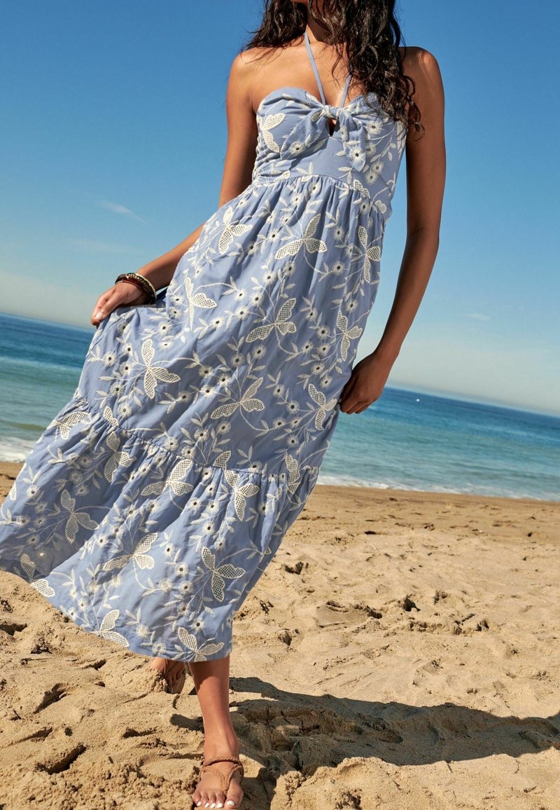 Femme en robe d'été bleu clair à fleurs et sandales, marchant sur une plage de sable avec un océan calme et un ciel dégagé en arrière-plan.