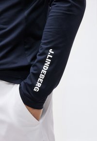 Άτομο με το χέρι στην τσέπη λευκού παντελονιού που φοράει μακρυμάνικο μπλε πουκάμισο με την επιγραφή "J.LINDEBERG" στο μανίκι.