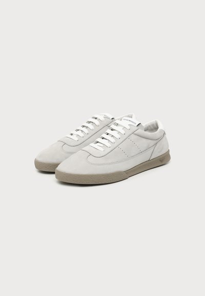Paire de baskets basses blanches avec des détails gris clair, des lacets blancs et des semelles en caoutchouc beige, posées sur un fond blanc uni.