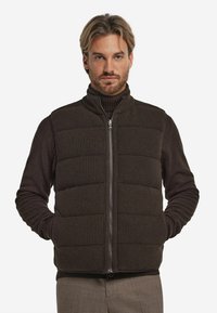 Braune Steppweste mit vertikaler Rippstruktur, Reißverschluss und hohem Kragen. Über einem dunklen Rollkragenpullover getragen und mit hellen Hosen kombiniert.