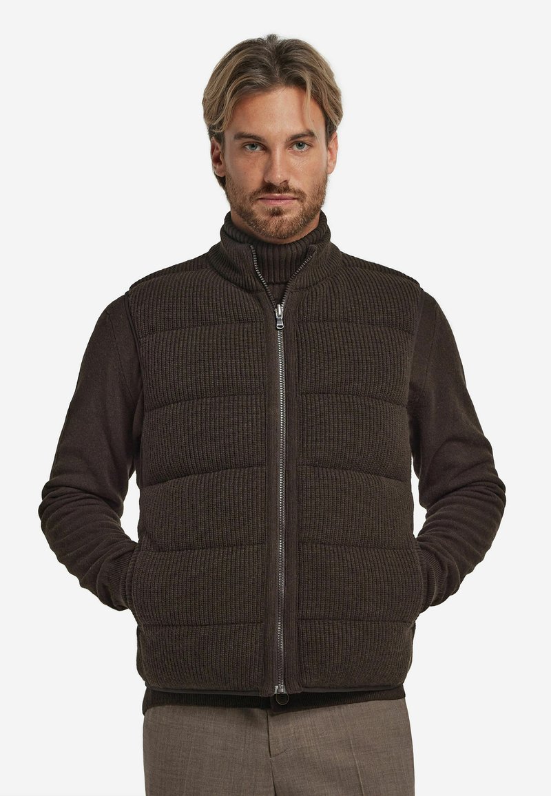 Braune Steppweste mit vertikaler Rippstruktur, Reißverschluss und hohem Kragen. Über einem dunklen Rollkragenpullover getragen und mit hellen Hosen kombiniert.