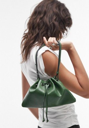 Vrouw met golvend bruin haar draagt een groen leren handtas met trekkoord over haar schouder, gekleed in een wit mouwloos topje.