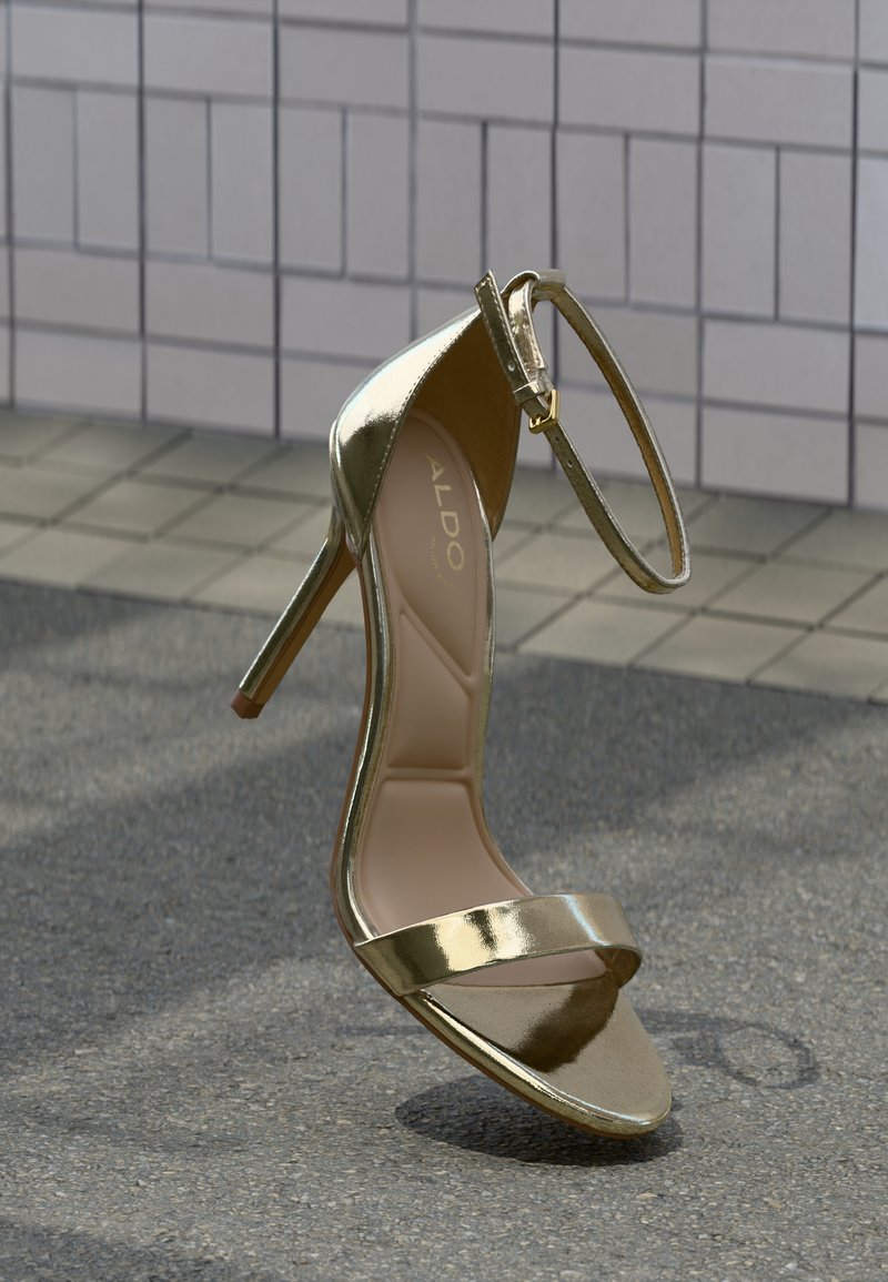 Sandale à talon haut dorée avec une finition métallique brillante, design à bout ouvert, fine bride à la cheville et semelle intérieure rembourrée. Nom de la marque visible à l'intérieur.