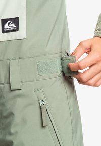 Groene, waterdichte sneeuwbroeken met een verstelbare taille van klittenband, een zijrits zak en een Quiksilver-logo. Glad oppervlak.