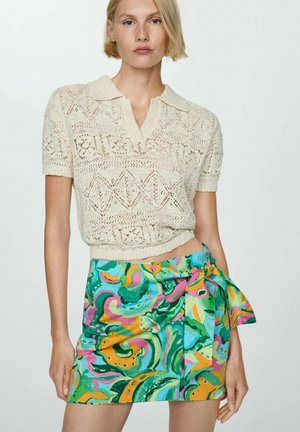 Femme portant un pull court à col en tricot crème à manches courtes et une mini-jupe en imprimé abstrait coloré avec un nœud sur le côté.