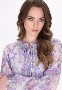 Bluză florală realizată dintr-un material ușor, transparent, cu modele în nuanțe de violet, roz și albastru. Decolteu cu volane și mâneci bufante. Detaliu cu șnur ajustabil.