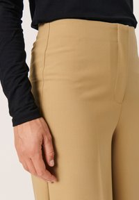 Beige-housut, jotka on valmistettu sileästä kankaasta, istuva malli, korkea vyötärö ja hienovarainen kiilto. Käsi lepää sivusaumassa.