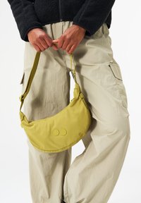 Gelbe Stoff-Schultertasche mit runder Form, ausgestattet mit zwei kreisförmigen Akzenten auf der Vorderseite und einem langen verstellbaren Tragegurt.