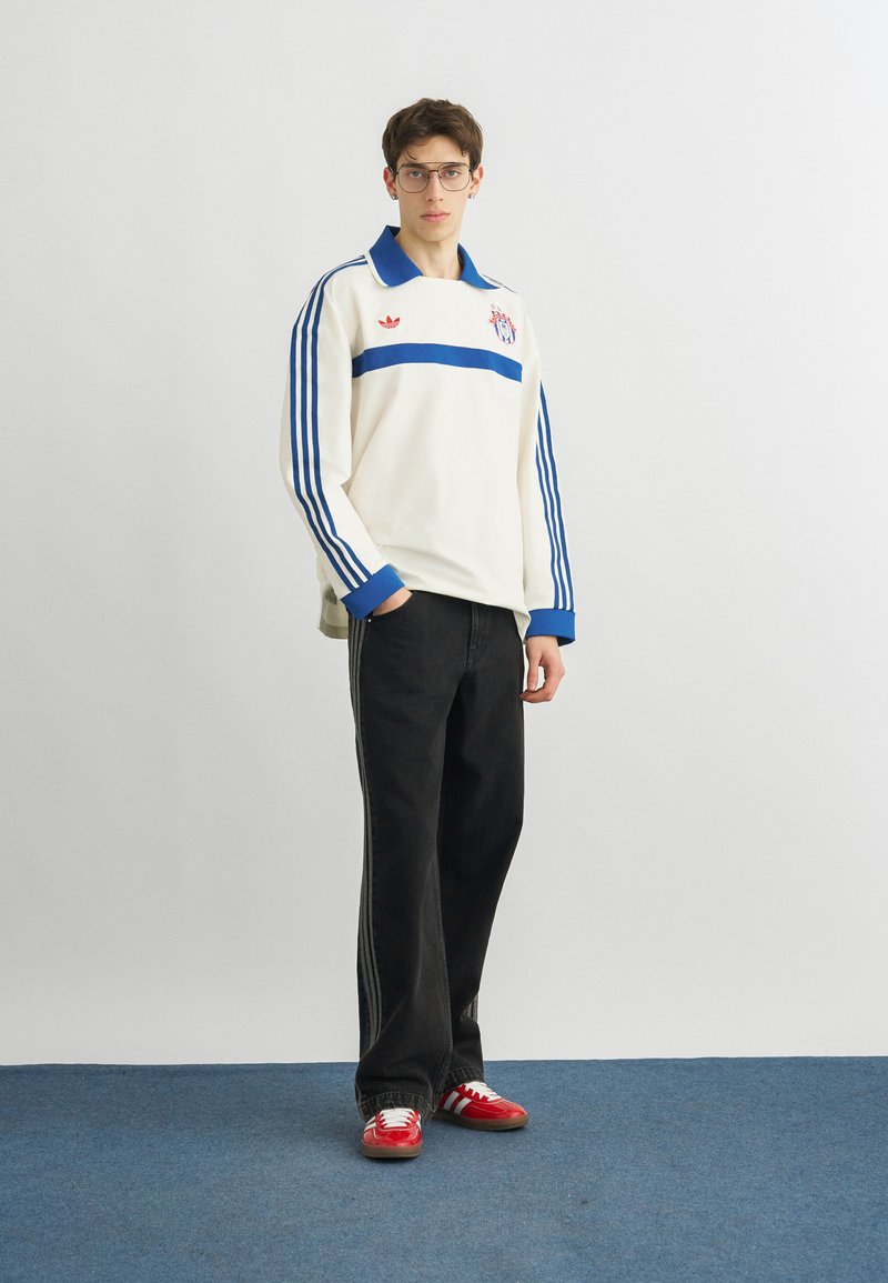 Giovane uomo con camicia bianca a maniche lunghe con strisce blu, pantaloni neri, sneakers rosse e occhiali, in piedi su un tappeto blu contro una parete bianca liscia.