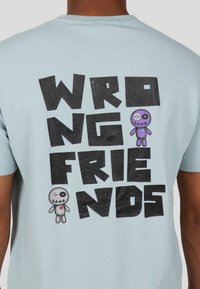 Vaaleansininen t-paita, jossa on takapainatus, jossa lukee "VÄÄRÄT YSTÄVÄT" paksuilla mustilla kirjaimilla. Kaksi sarjakuvamaista pehmolelua, harmaa ja violetin värinen, on esillä.