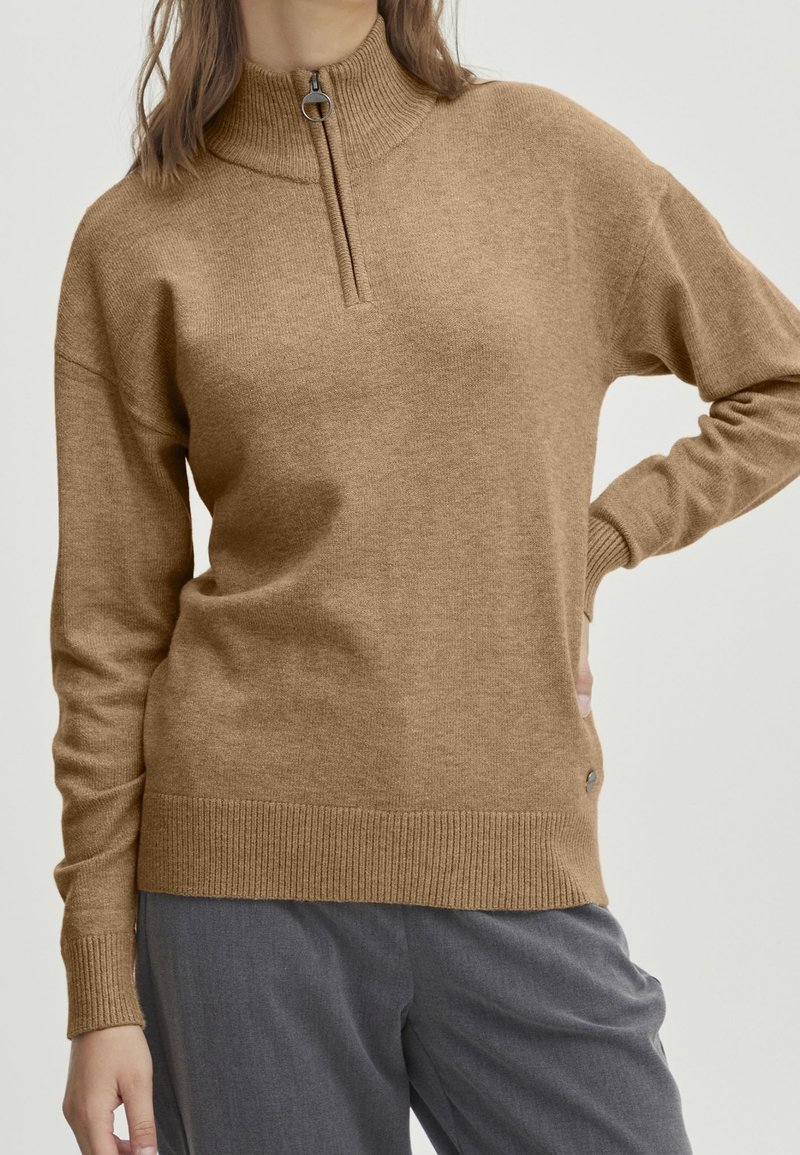 Femme portant un pull en maille marron chameau avec un col court zippé et un pantalon gris ample, debout avec une main sur la hanche.