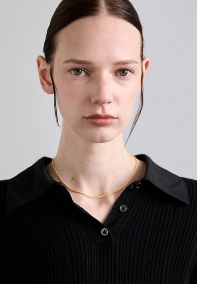 Camisa de rayas negra con cuello, con botones negros, acompañada de un delgado collar dorado trenzado. Fondo neutro.