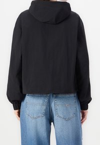 Czarna, krótka kurtka z kapturem, z elastycznymi mankietami i lekką teksturą, połączona z wysokimi, wypłukanymi niebieskimi dżinsami.