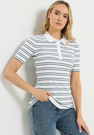 Blondynka w białej koszulce polo z czarnymi paskami i niebieskich dżinsach, stojąca przy białej ścianie, z jedną ręką w kieszeni.