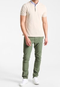 A man stands in a neutral background, wearing a light beige polo shirt, green pants, and white sneakers, arms relaxed at his sides.