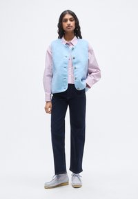 Ανοιχτό μπλε γιλέκο από φλις με πέντε κουμπιά, φορεμένο πάνω από ένα ροζ ριγέ πουκάμισο. Συνδυασμένο με σκούρο μπλε τζιν και γκρι παπούτσια με κορδόνια.