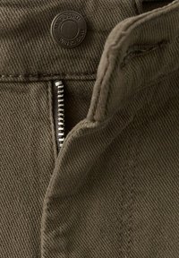 Nahaufnahme von dunkelolivgrünem Denimstoff mit einem Knopf mit der Aufschrift "MNG DENIM" und einem teilweise geöffneten Metallreißverschluss.