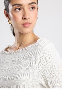 White, smocked top with a wide, ruffled neckline. The fabric is textured, soft, and features subtle horizontal patterns. Gold hoop earrings accent the look.