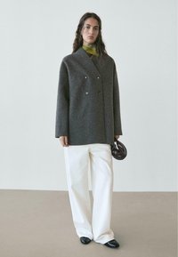 Cappotto doppiopetto in lana grigia con grandi bottoni, abbinato a pantaloni crema a gamba larga e una sciarpa verde, con in mano una borsa marrone scuro.