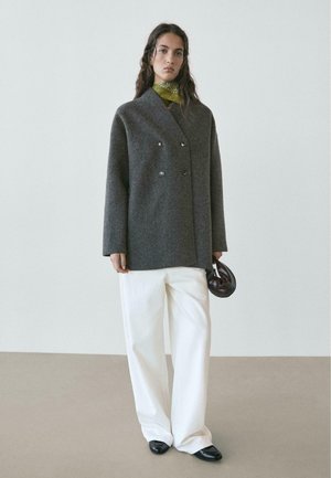 Cappotto doppiopetto in lana grigia con grandi bottoni, abbinato a pantaloni crema a gamba larga e una sciarpa verde, con in mano una borsa marrone scuro.