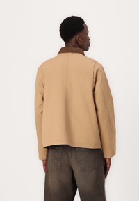 Tan jacket with a brown corduroy collar, featuring a relaxed fit and visible stitching. Worn with dark gray jeans, seen from the back.
