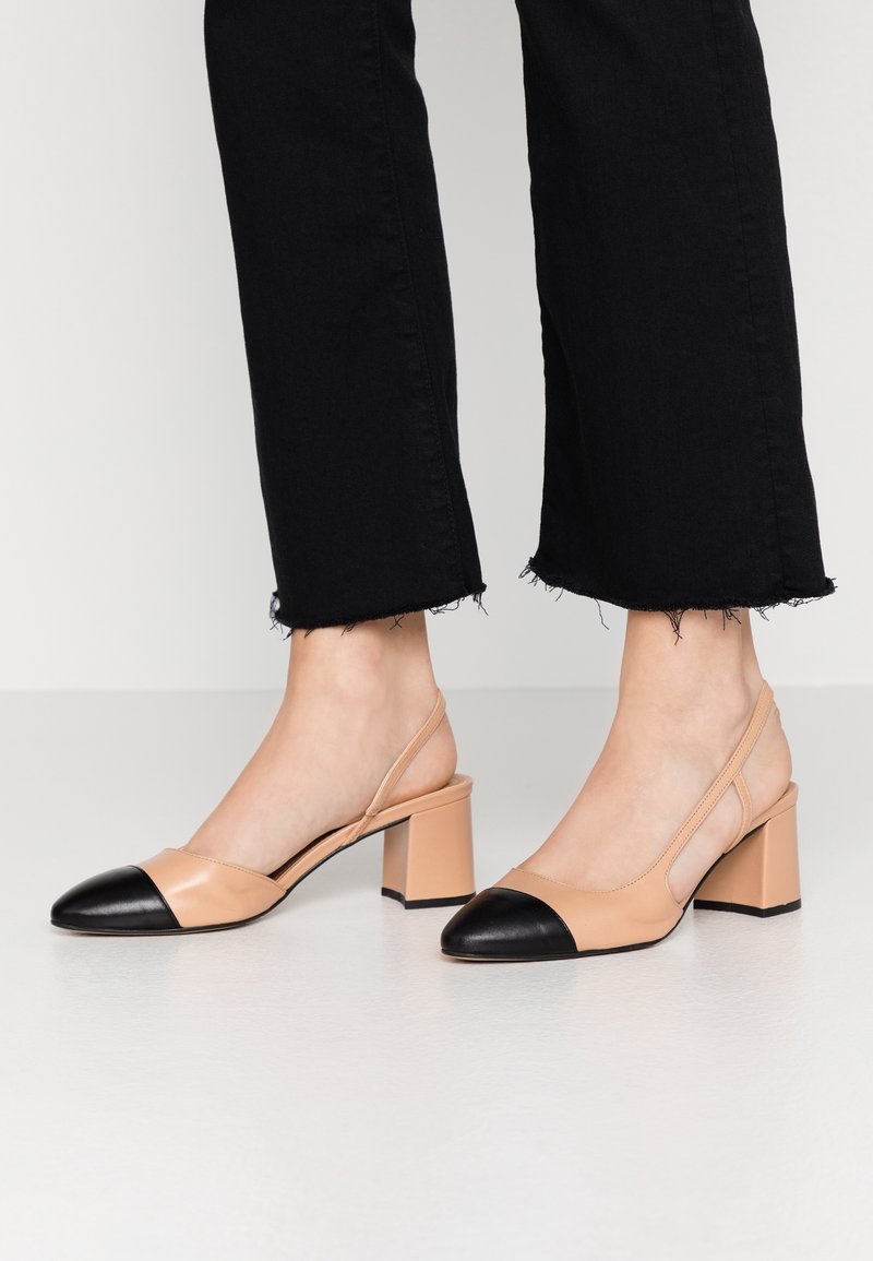 Escarpins à talons blocs beige avec bouts pointus noirs portés avec un pantalon court noir à ourlet effiloché, sur un sol clair.