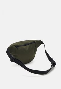 Sac banane en tissu vert olive avec une sangle noire réglable et une poche zippée, présentant une forme arrondie et une texture lisse.