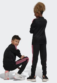 Schwarze Sportanzüge mit pinken Streifen, aus flexiblem Material. Enthält kontrastierende Sneakers: weiß mit pinken Akzenten und schwarz mit orange.