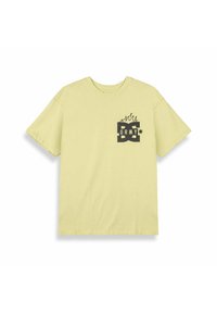 T-shirt di cotone gialla con un design grafico nero che include le lettere "DC" e "GRMY", con accenti di fiamma nell'area del petto sinistro.