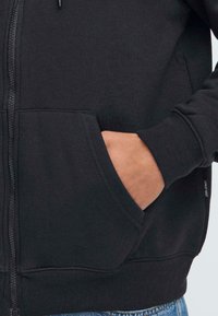 Svart zip-up hettegenser med kengurulomme, ribbestrikkede mansjetter og hem. Myk stofftekstur med subtil merkevaredetalj på siden.
