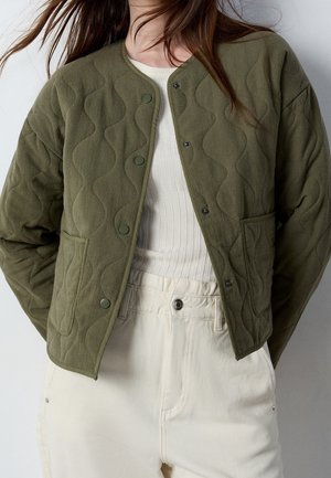 Femme portant une veste matelassée vert olive avec boutons-pression, superposée à un haut blanc côtelé et un pantalon taille haute blanc cassé.