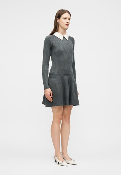 Femme debout portant une robe en maille côtelée grise à manches longues avec un col pointu blanc et des chaussures blanches à talons bas, sur un fond uni.