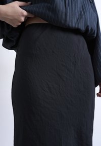 Χέρι σηκώνει ριγωτό μπλε πουκάμισο, αποκαλύπτοντας γυμνό δέρμα πάνω από μαύρη υφασμάτινη φούστα με ψηλή μέση, με φόντο απλό ανοιχτόχρωμο.