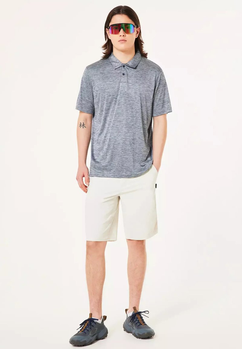 Joven con polo gris, pantalones cortos blancos, zapatillas oscuras y gafas de sol reflectantes con diseño de arcoíris, de pie con una mano en el bolsillo.