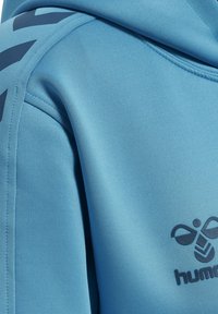 Close-up van een blauwe hoodie met zicht op de schoudernaden, gedeeltelijke mouw en een donker logo met bij en de tekst "hummel" op de borst.