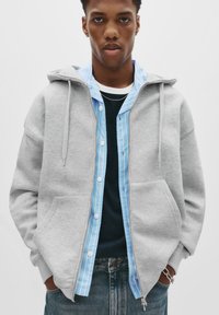 Young man wearing a light gray zip-up hoodie over a blue striped shirt and dark t-shirt, with hands in jacket pockets and denim jeans.