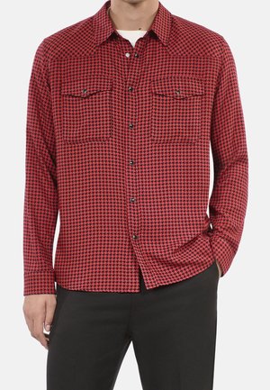 Homme portant une chemise à boutons rouge et noire à motif pied-de-poule avec des poches poitrine, associée à un pantalon noir, main dans la poche.