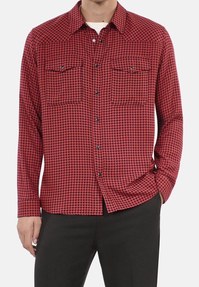 Homme portant une chemise à boutons rouge et noire à motif pied-de-poule avec des poches poitrine, associée à un pantalon noir, main dans la poche.