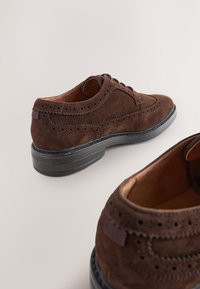 Chaussures en daim marron avec détails brogue, bout arrondi, semelle extérieure plate noire et lacets. L'intérieur a une texture en cuir et une couleur claire.
